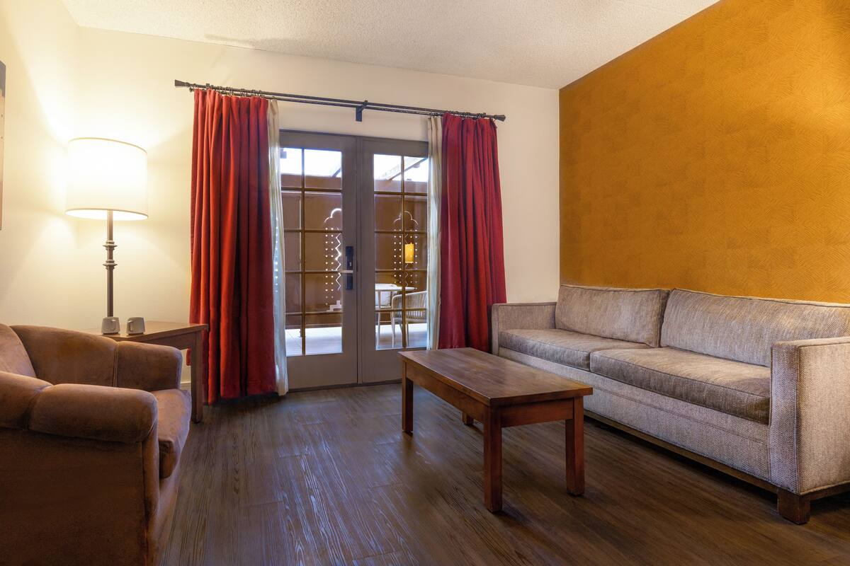 Una sala de estar con sofá, una mesa de centro de madera, una lámpara y vistas a través de un ventanal con cortinas rojas.