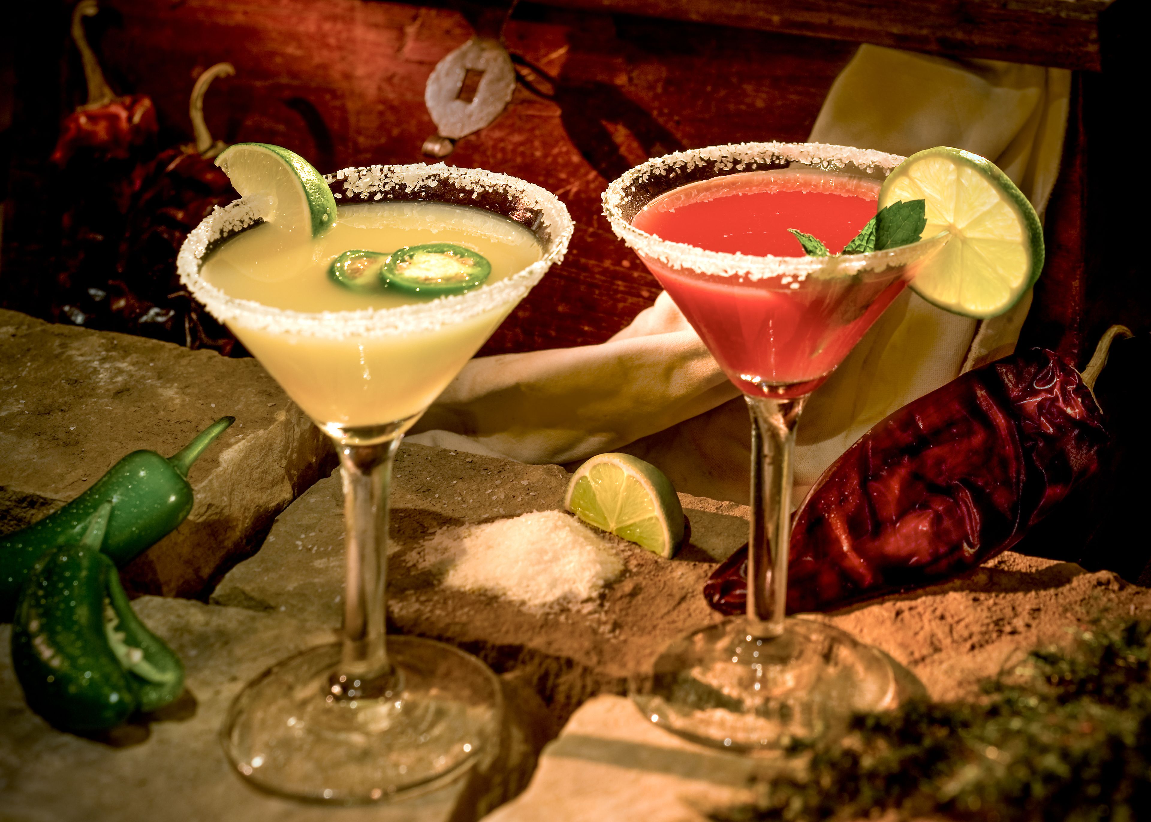 Two cocktails with lime slices and a salt rim, surrounded by green chili peppers and a rustic wooden background.