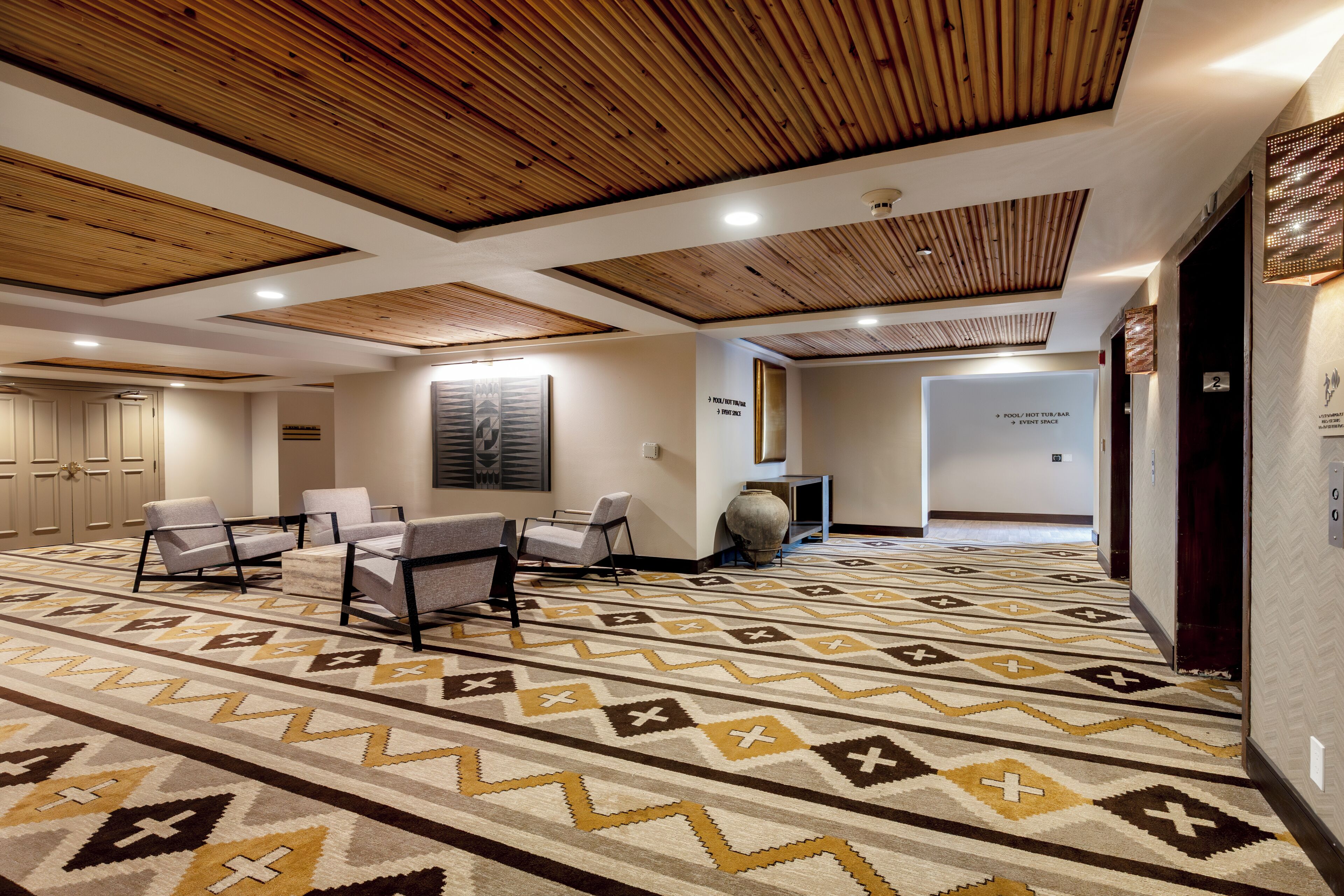 A hotel lobby with a patterned carpet, wooden ceiling, seating area, and directional signs.