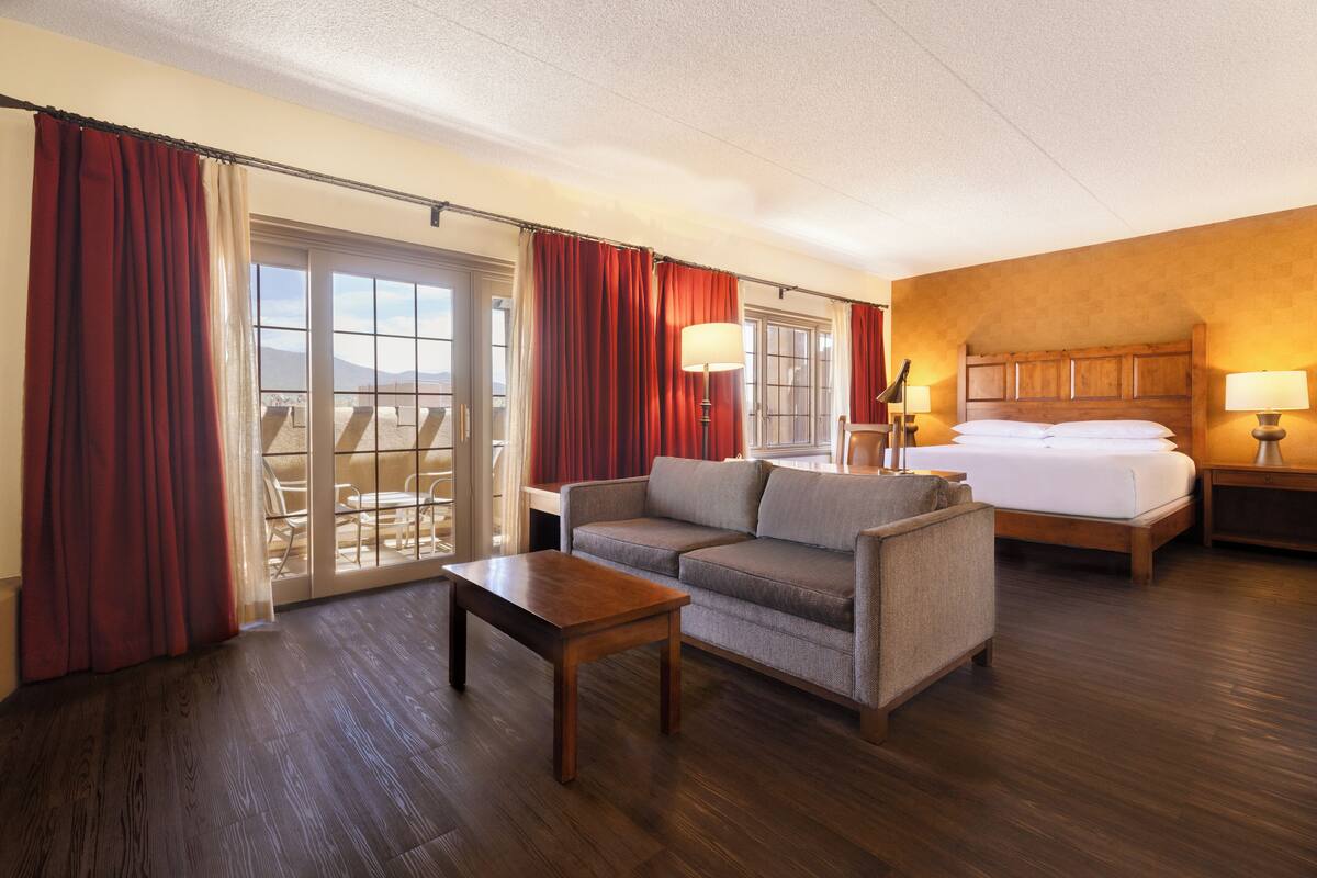 Habitación de hotel con sofá, cama, mesa de madera y balcón con cortinas rojas.
