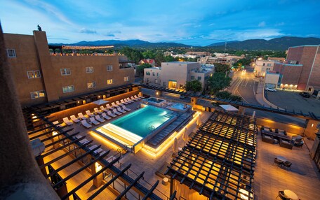 A rooftop pool area with lounge chairs and a hot tub, surrounded by buildings and a view of the mountains.