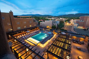 Un área de piscina en la azotea con sillones de descanso y un jacuzzi, rodeada de edificios y vistas a las montañas.