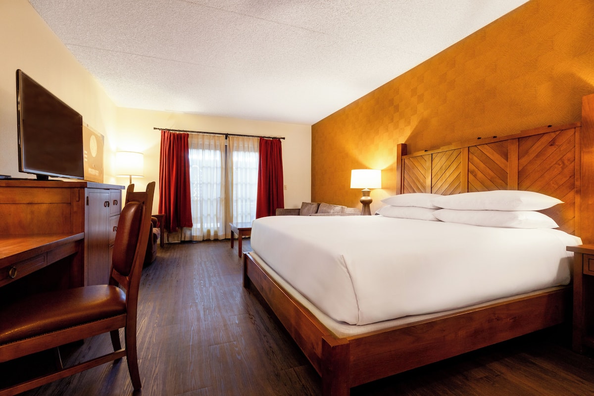 Habitación de hotel con una cama de madera, un escritorio con una silla, un televisor y una ventana con cortinas.
