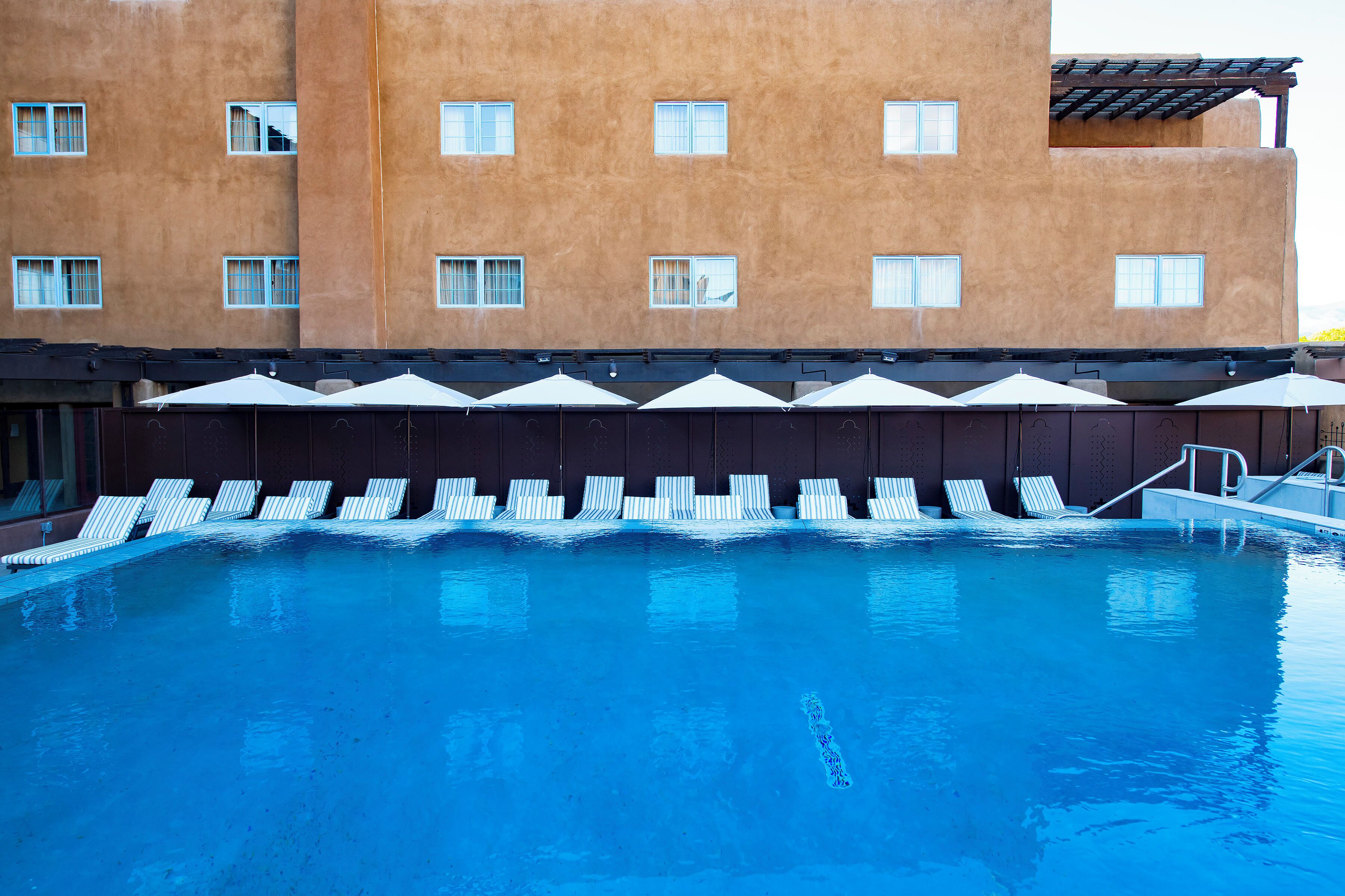 A swimming pool with lounge chairs and umbrellas, a building with multiple windows, and a clear sky.
