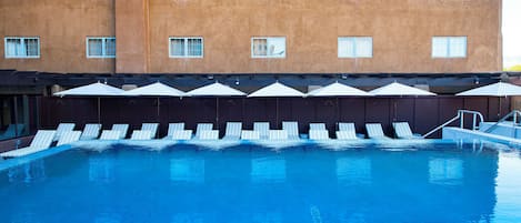 A swimming pool with lounge chairs and umbrellas, a building with multiple windows, and a clear sky.