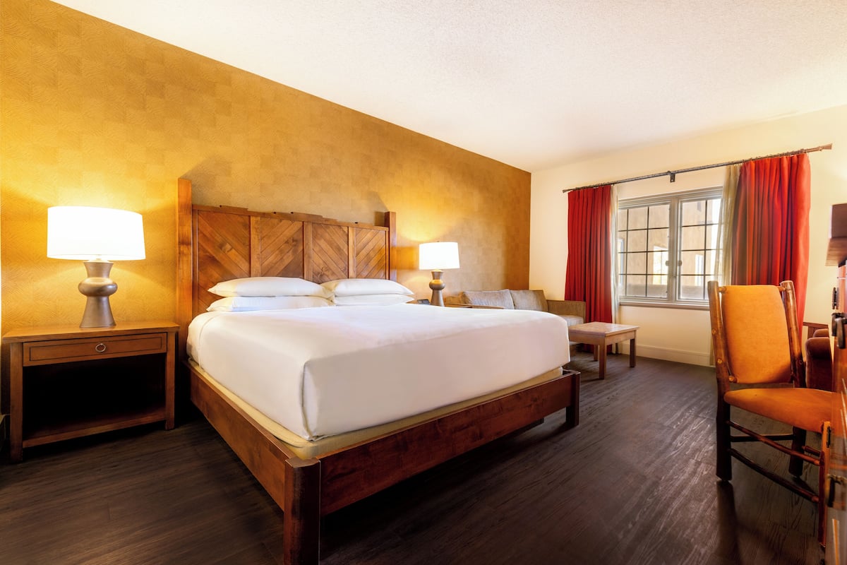 Habitación de hotel con una cama de madera, dos mesitas de noche con lámparas, una silla, una pequeña mesa y un ventanal con cortinas.