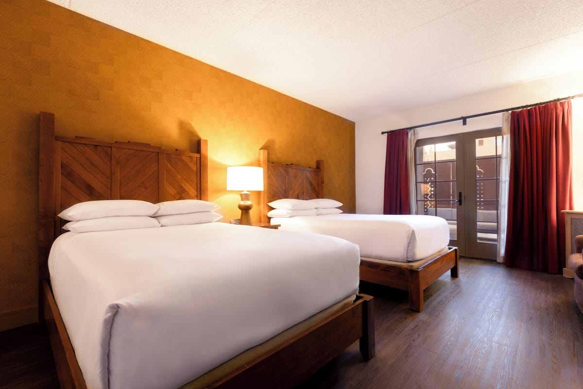 Habitación de hotel con dos camas, cabeceros de madera, una mesita de noche con lámpara y una puerta corrediza de vidrio con cortinas rojas.