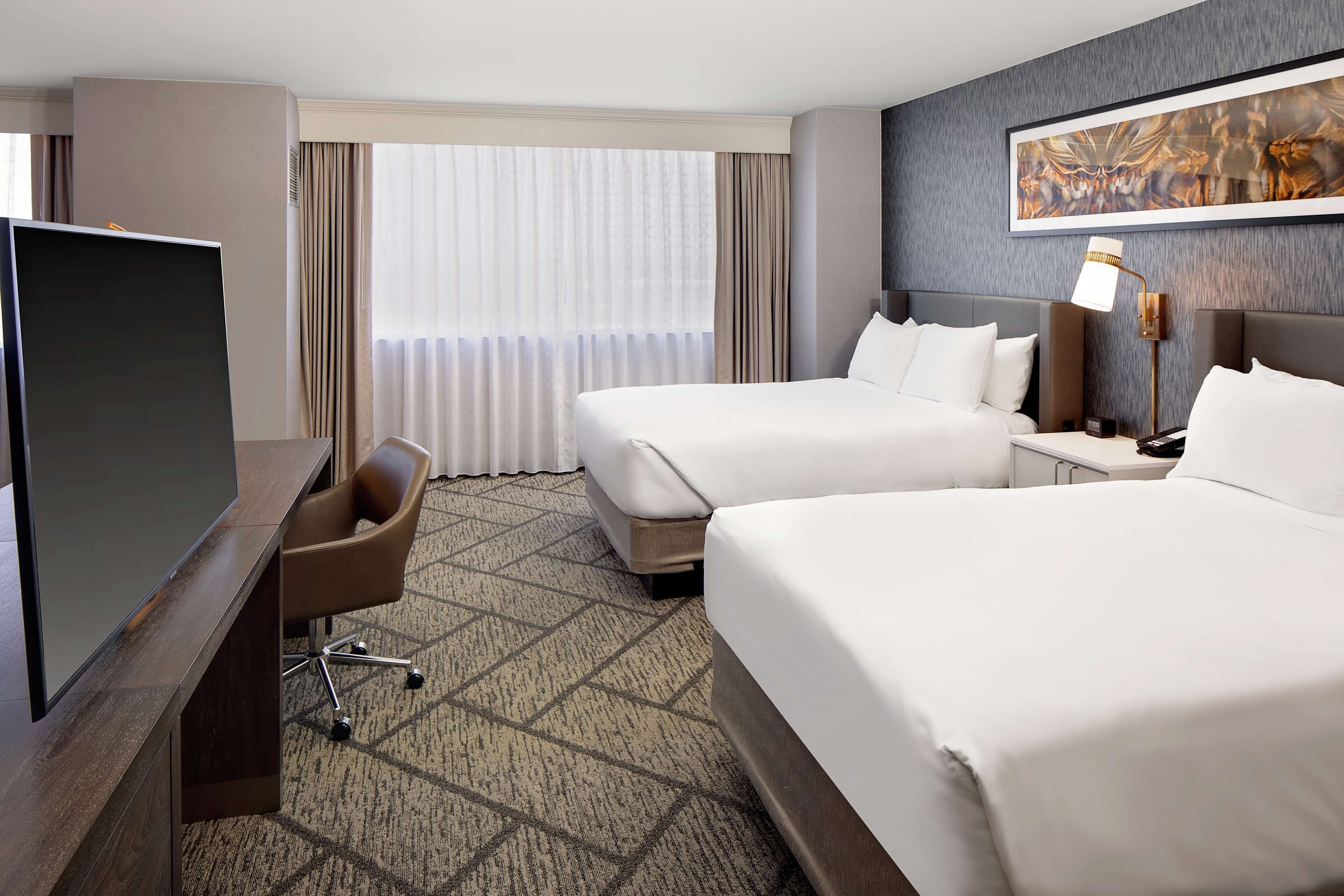 A hotel room with two beds, a desk with a flat-screen TV, a chair, and a large window with curtains.