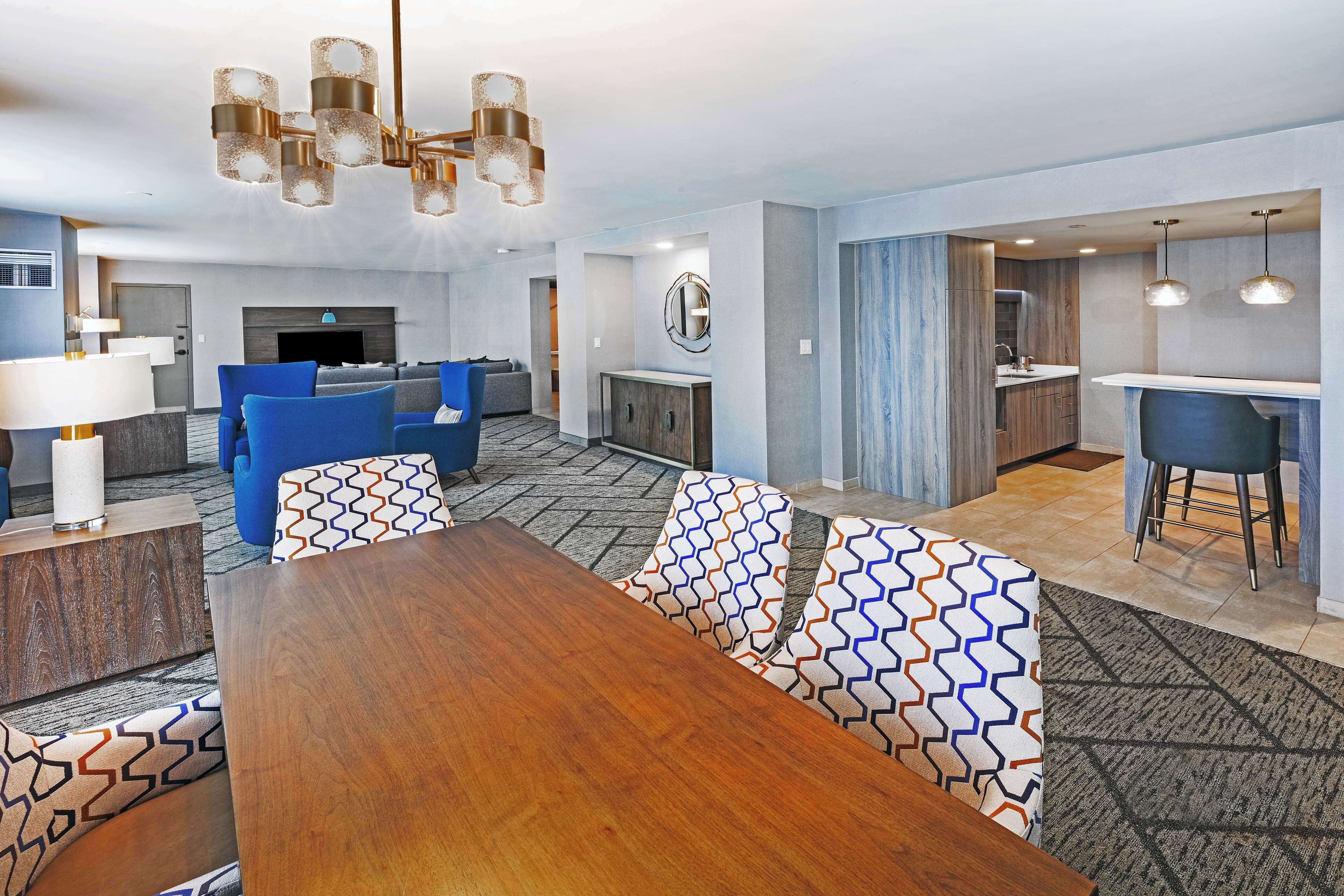 A modern hotel room with a wooden dining table, blue sofas, a kitchen area, and a living room with a fireplace.