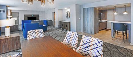 A modern hotel room with a wooden dining table, blue sofas, a kitchen area, and a living room with a fireplace.