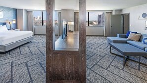 Habitación de hotel con una cama grande, un sofá, una mesa de centro, un televisor y vistas de la ciudad a través de amplios ventanales.