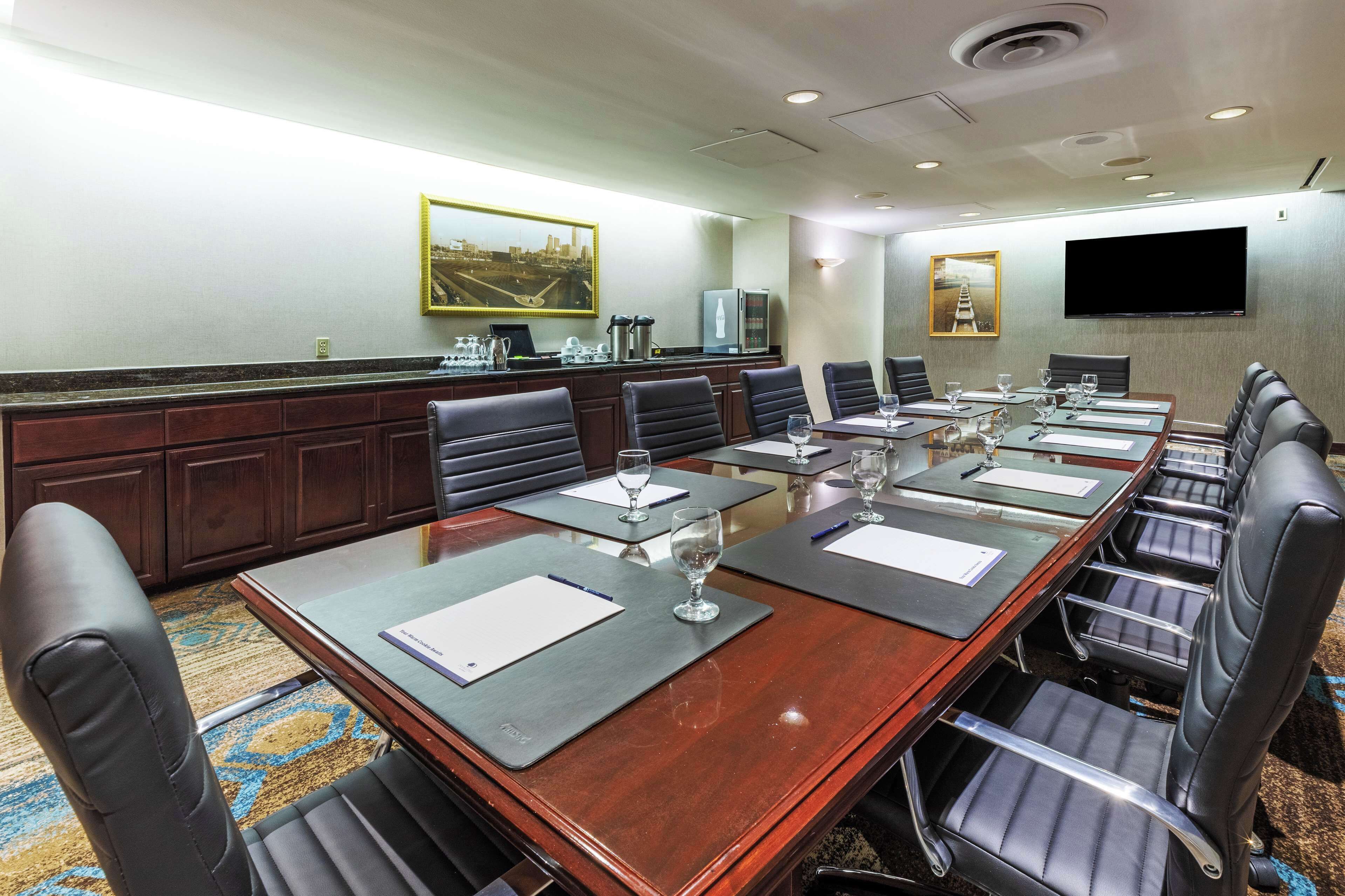 A conference room with a long table, multiple chairs, a flat-screen TV, and a kitchen area with a refrigerator and coffee maker.