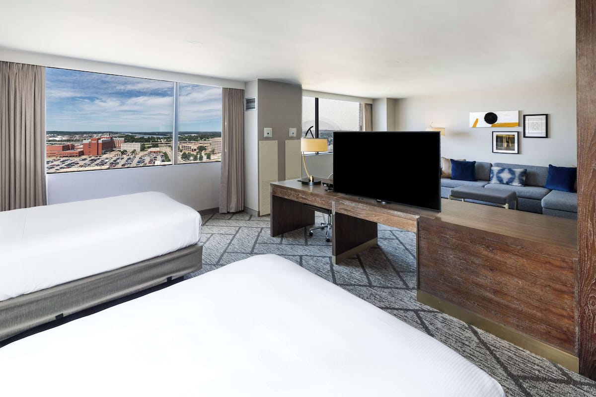 Ett hotellrum med två sängar, ett skrivbord med en tv, en soffa och ett stort fönster med utsikt över staden.