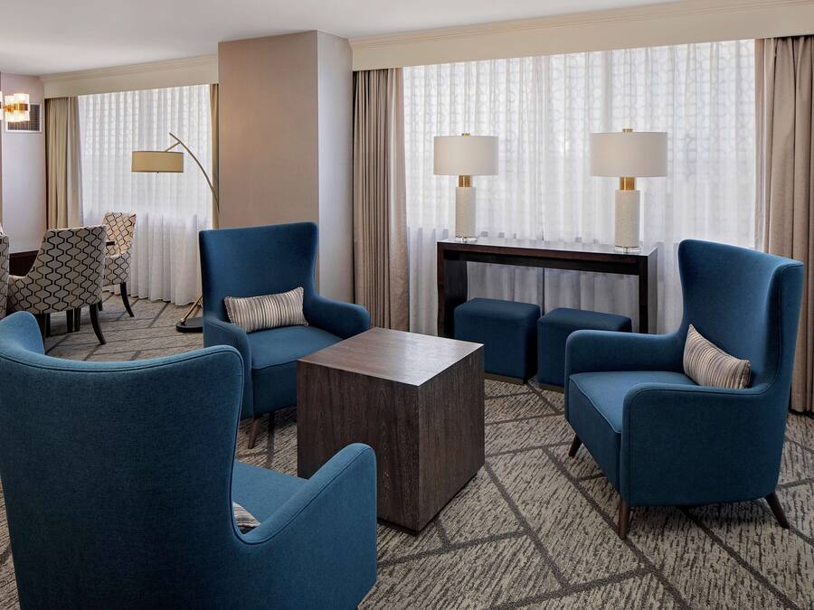 Een moderne hotelkamer met blauwe fauteuils, een donkerhouten salontafel en een kamerbreed tapijt met dessin.