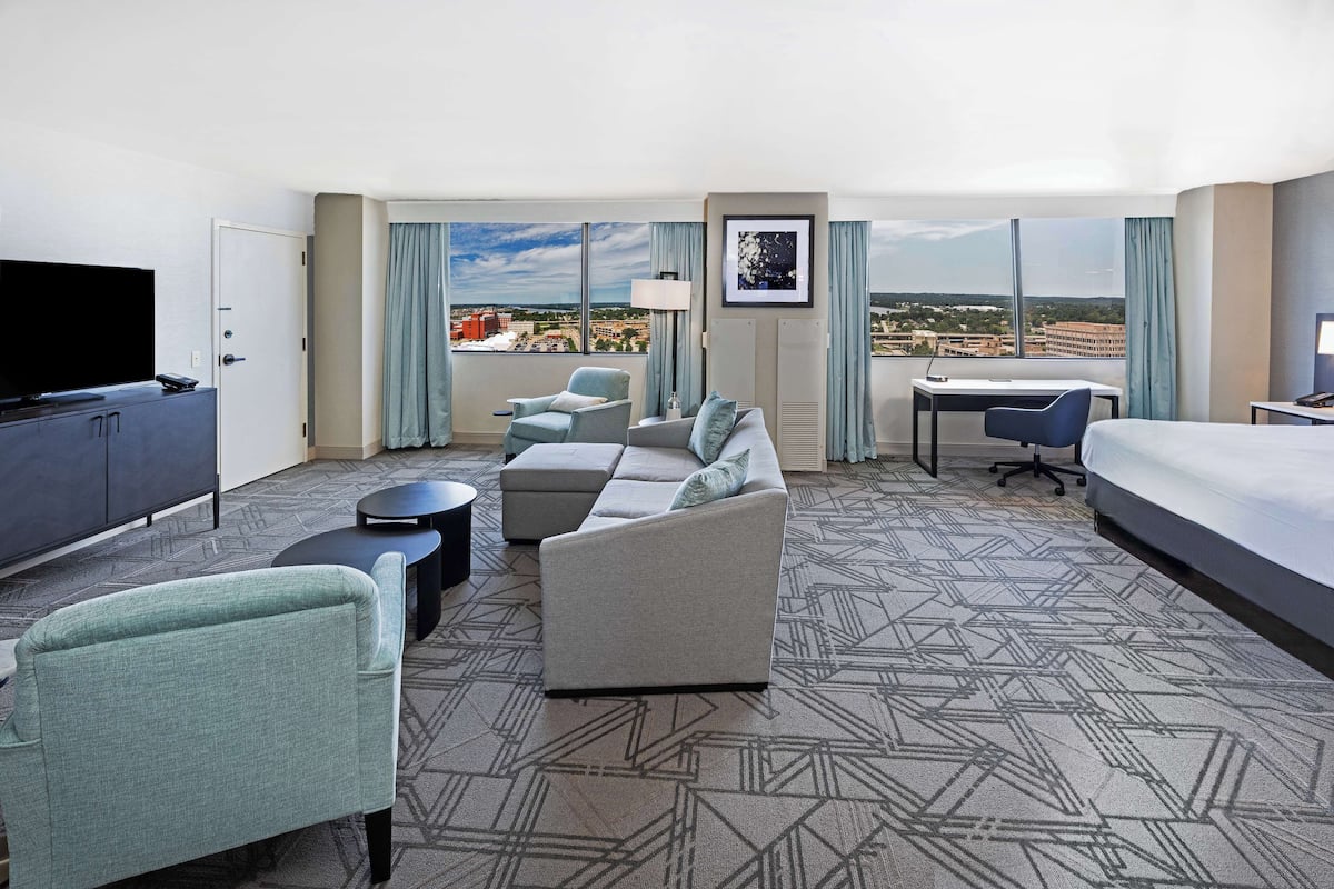 Ett hotellrum med en stor säng, en soffa, ett skrivbord, en TV och utsikt över staden.