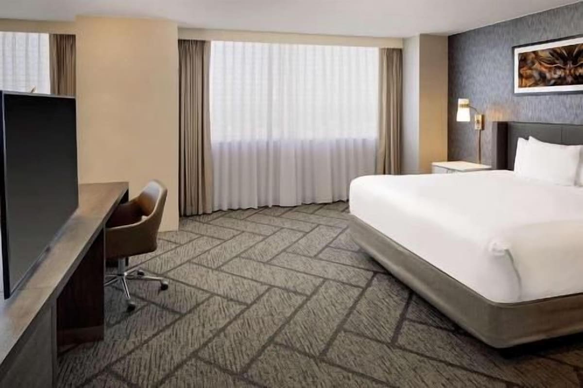 Une chambre d’hôtel avec un grand lit, un bureau, une chaise, une télévision et une fenêtre avec des rideaux.