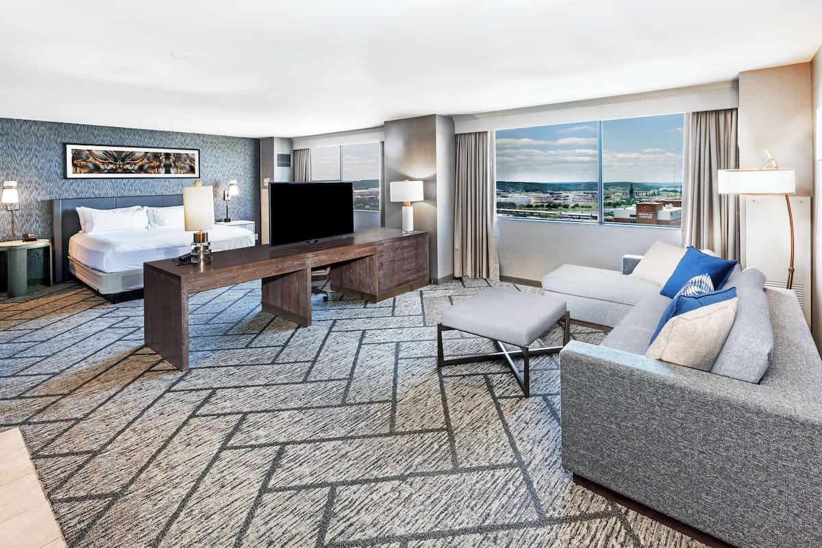 Ett hotellrum med en säng, ett skrivbord med en tv, en soffa och ett stort fönster med utsikt över staden.
