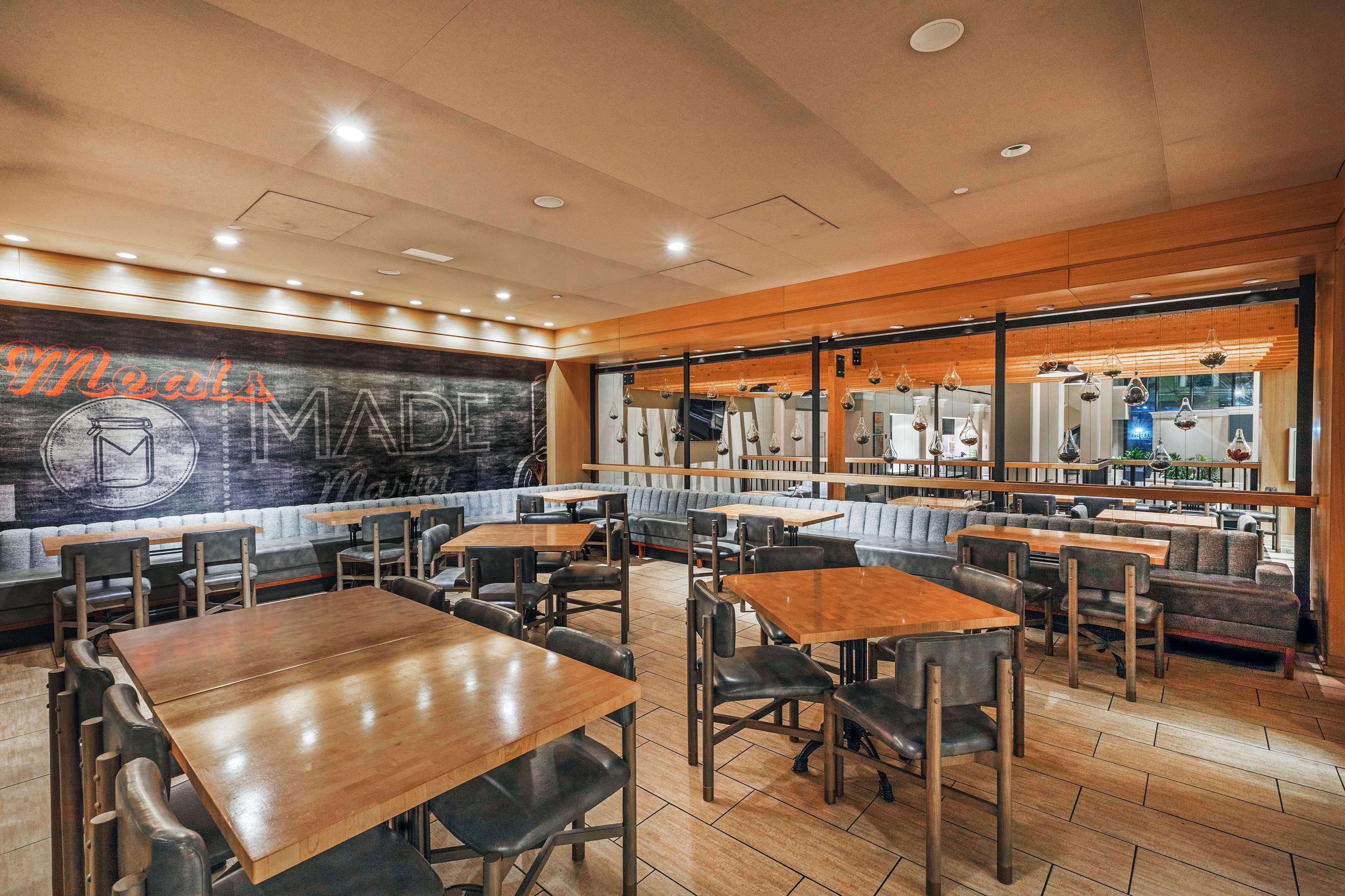 A restaurant with wooden tables and chairs, a large mural on the wall, and a view of the kitchen through the windows.
