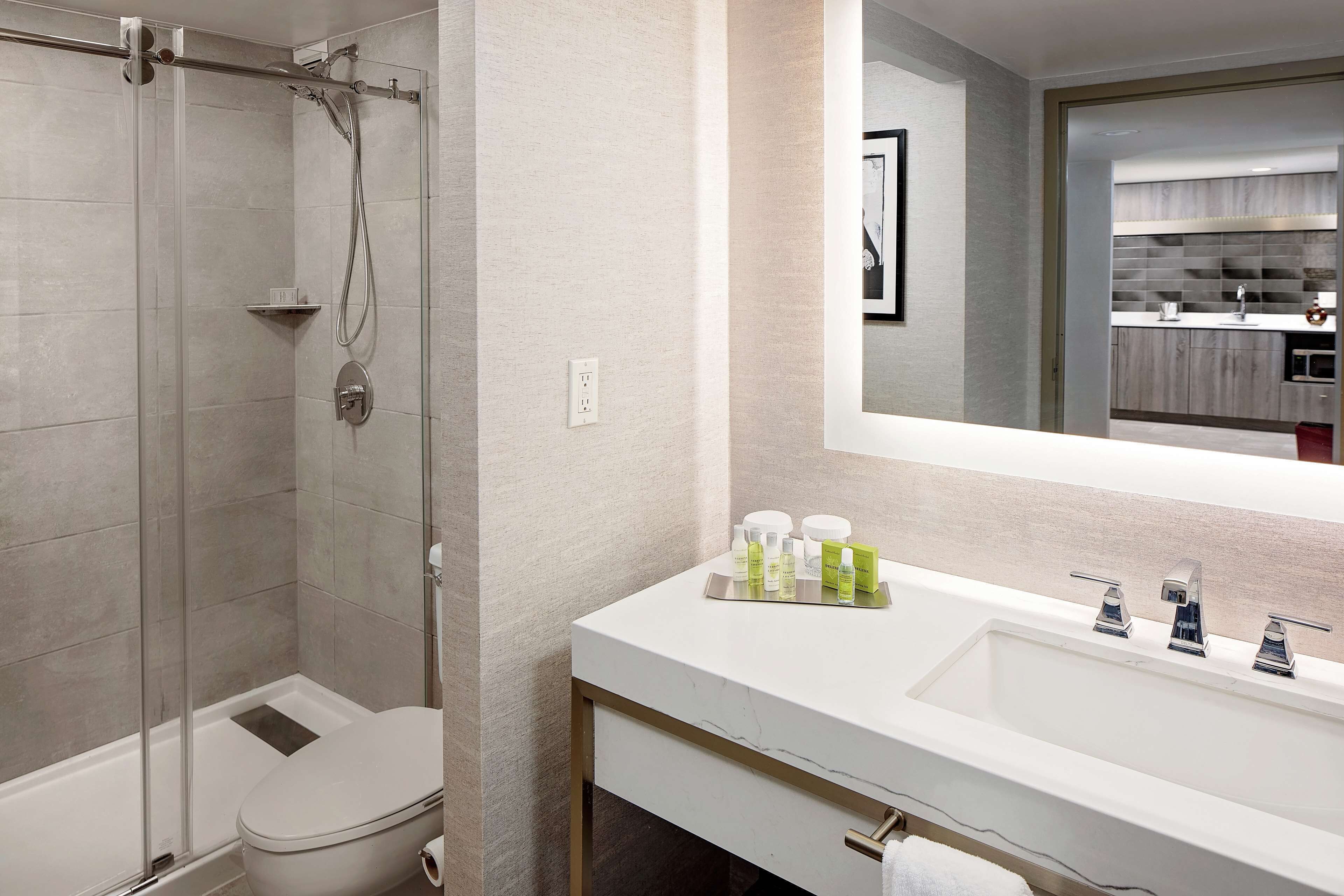 A modern bathroom with a glass-enclosed shower, a white sink with a marble countertop, and a large mirror reflecting a kitchen area.