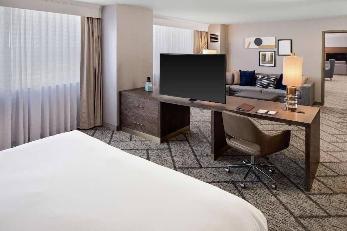 Ett hotellrum med en säng, ett skrivbord med en platt-TV, en stol, en lampa och en soffa.