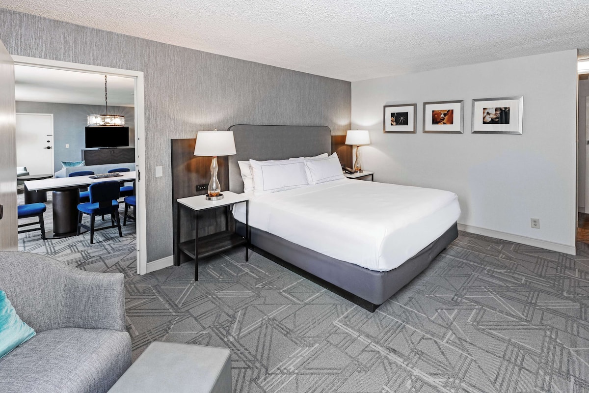 Ett hotellrum med en stor säng, två sängbord med lampor, ett skrivbord med stolar och en matplats som syns genom en öppen dörr.