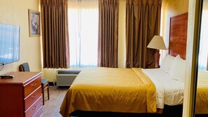 A hotel room with a bed, a desk with a chair, a television, and a window with curtains. - Coachman's Inn (Magnolia)