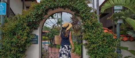 A woman in a hat and a dog walking through an arched entrance with greenery.