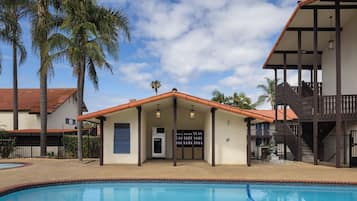 Uma área na beira da piscina com um edifício que possui um alpendre coberto, uma fonte de água e uma máquina de vendas.