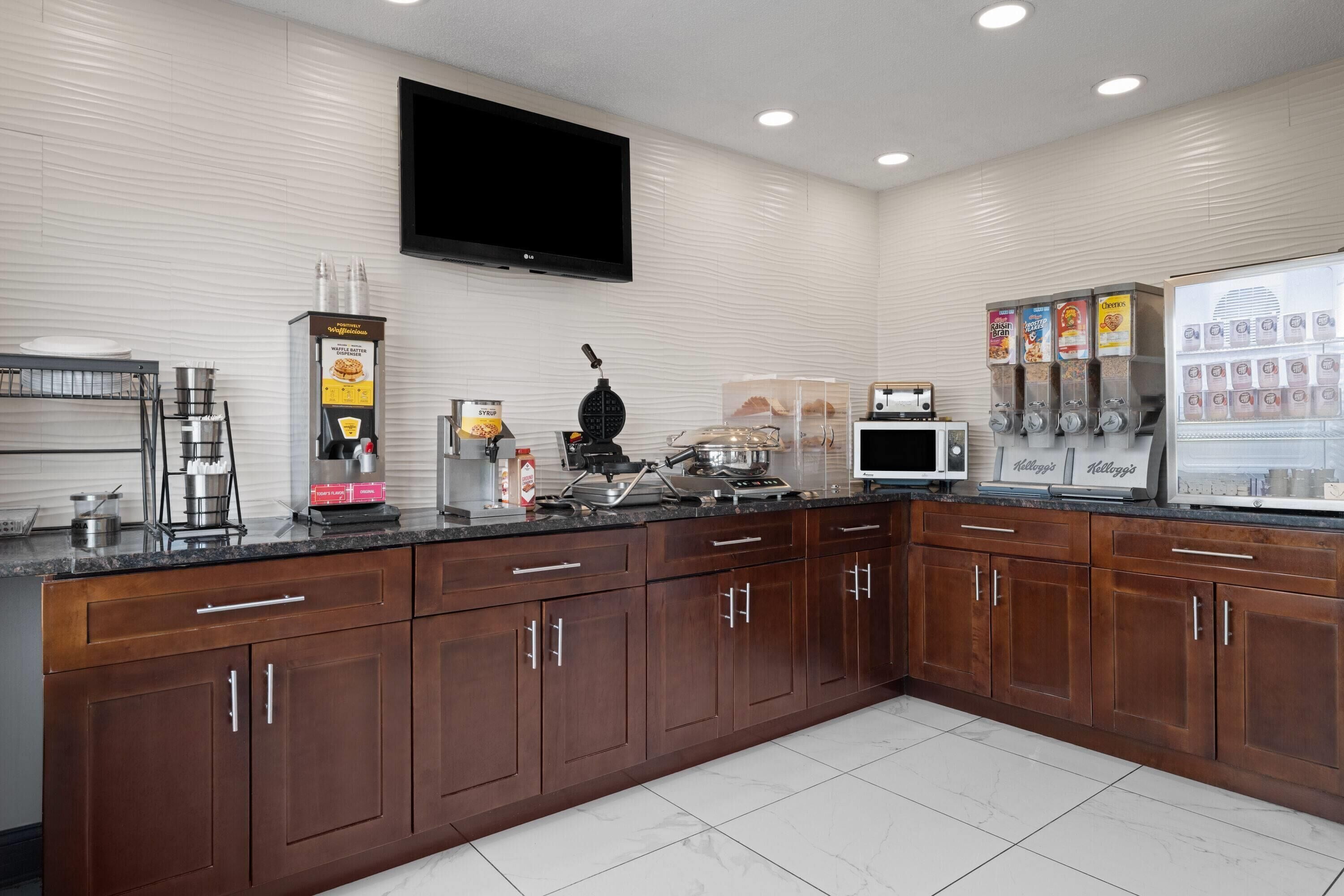 A kitchen with a flat-screen TV, coffee machines, a microwave, and a refrigerator stocked with beverages.