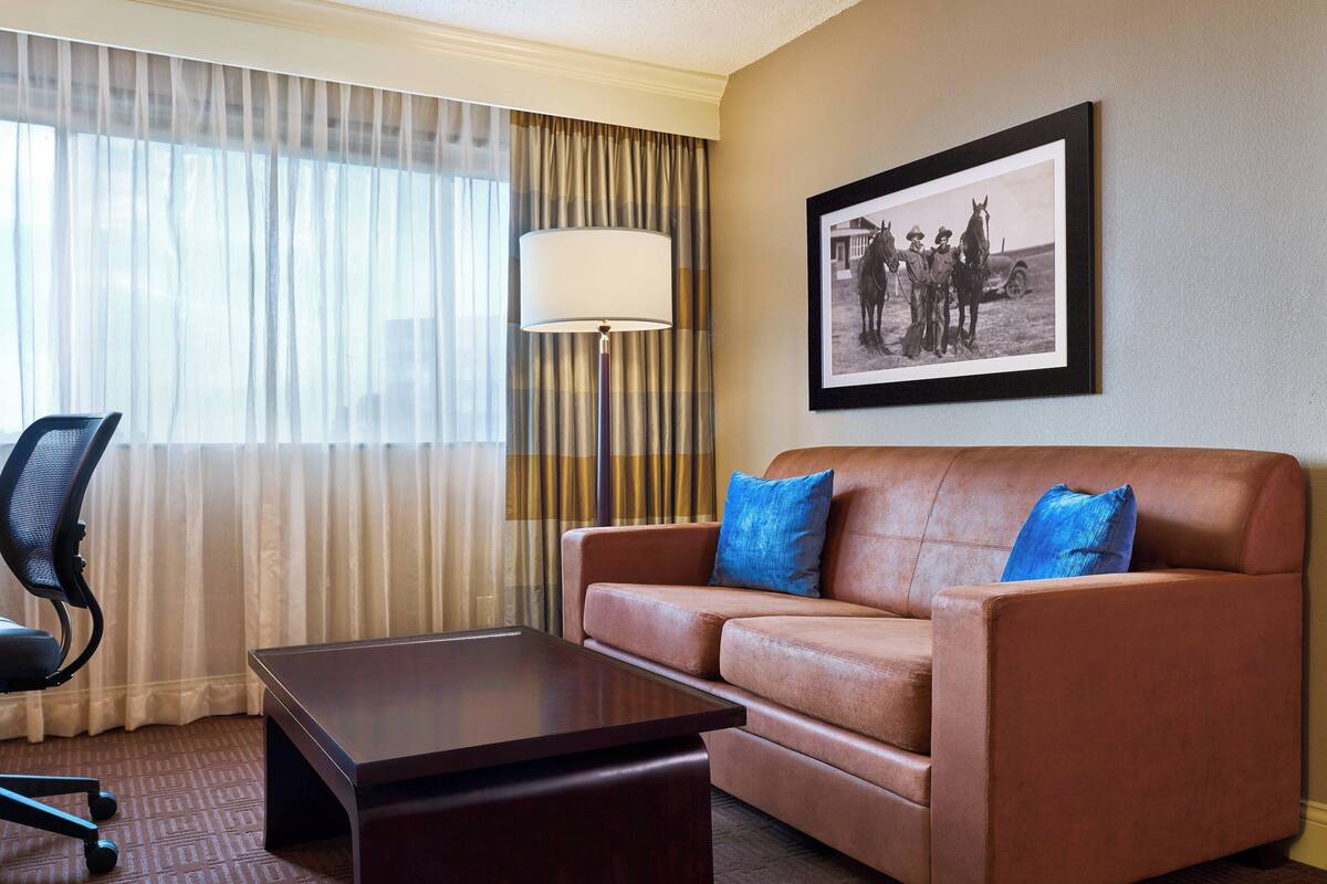 A hotel room with a brown leather sofa, a wooden coffee table, a black office chair, and a large window with sheer curtains.