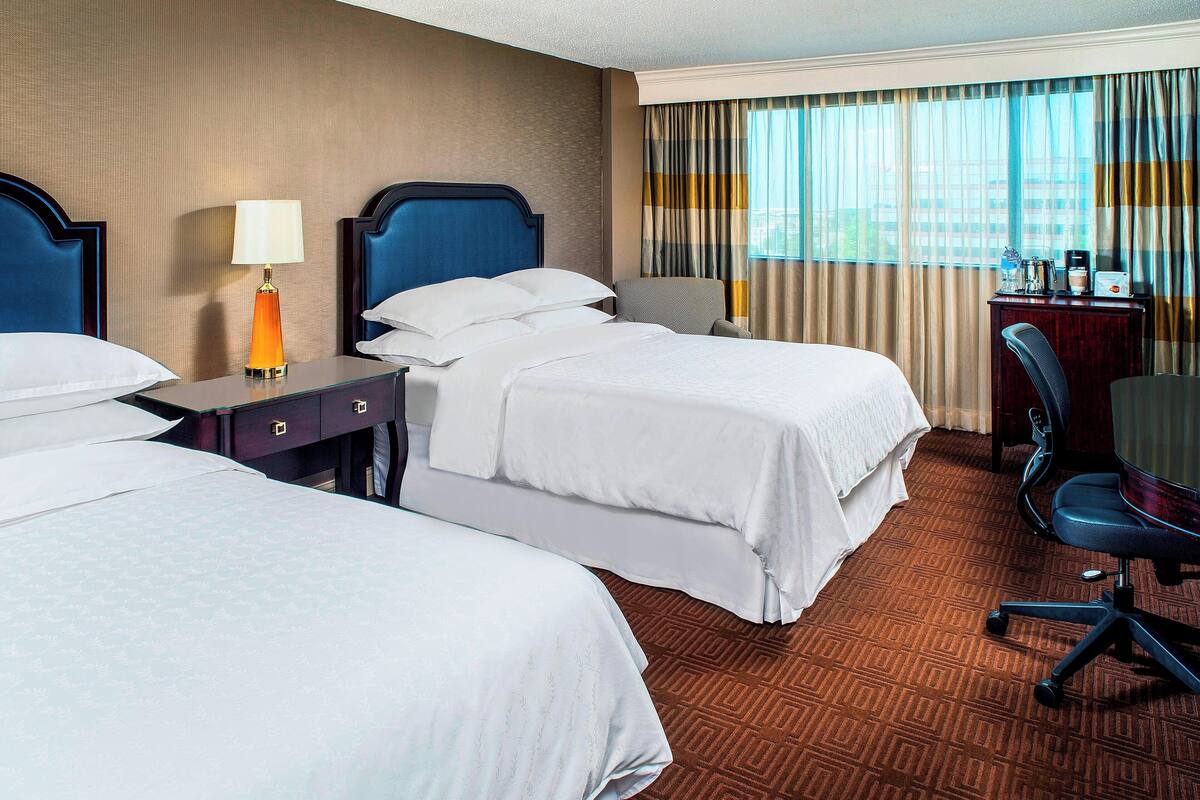 A hotel room with two beds, a desk with a lamp, a chair, and a view of the city through large windows.
