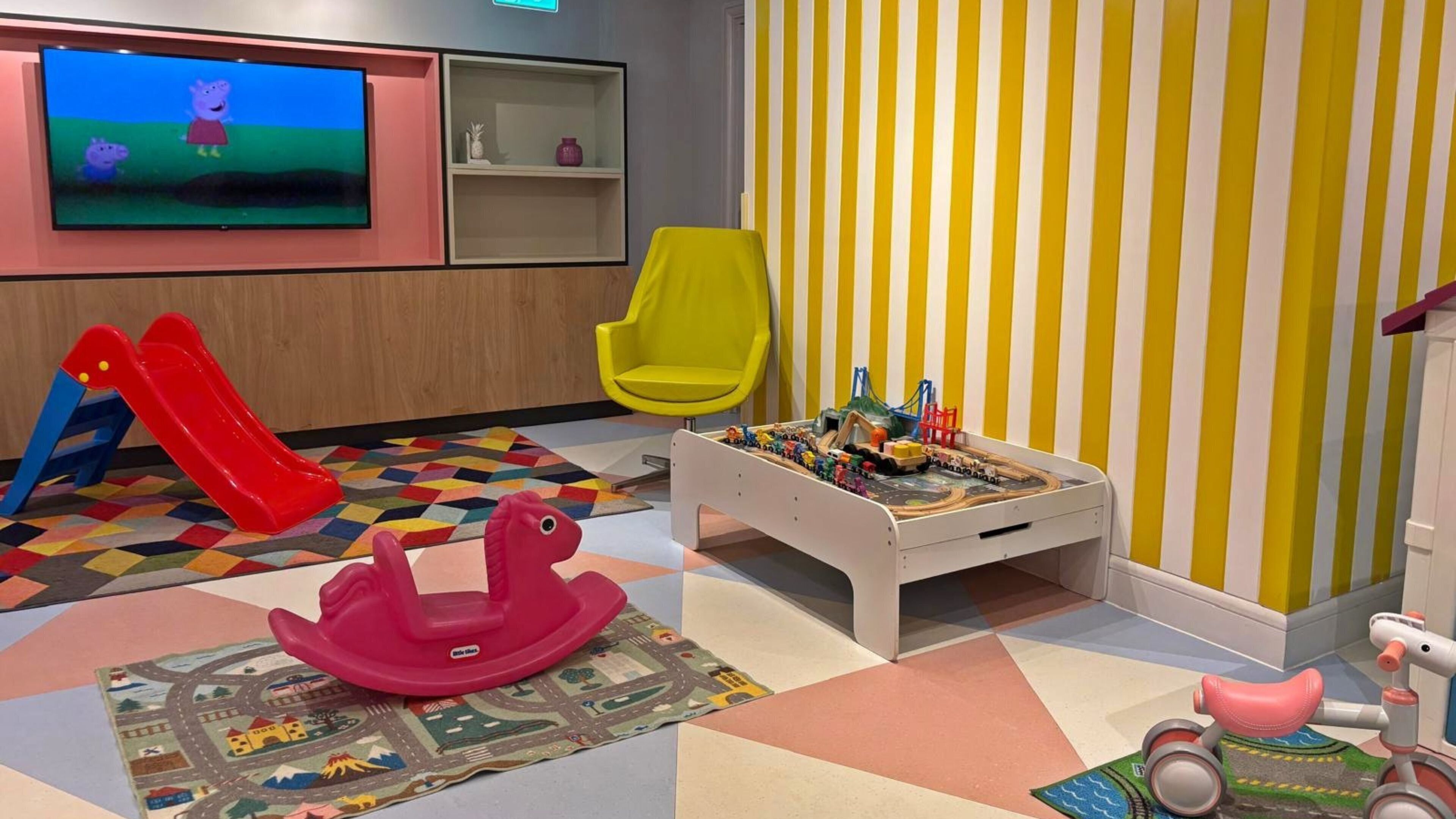 A children's play area with a TV, a yellow chair, a play table with toys, a red slide, and a pink rocking horse.