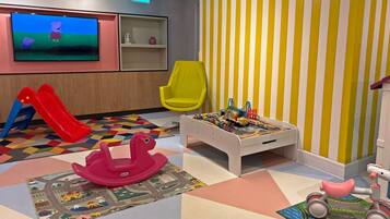 A children's play area with a TV, a yellow chair, a play table with toys, a red slide, and a pink rocking horse.