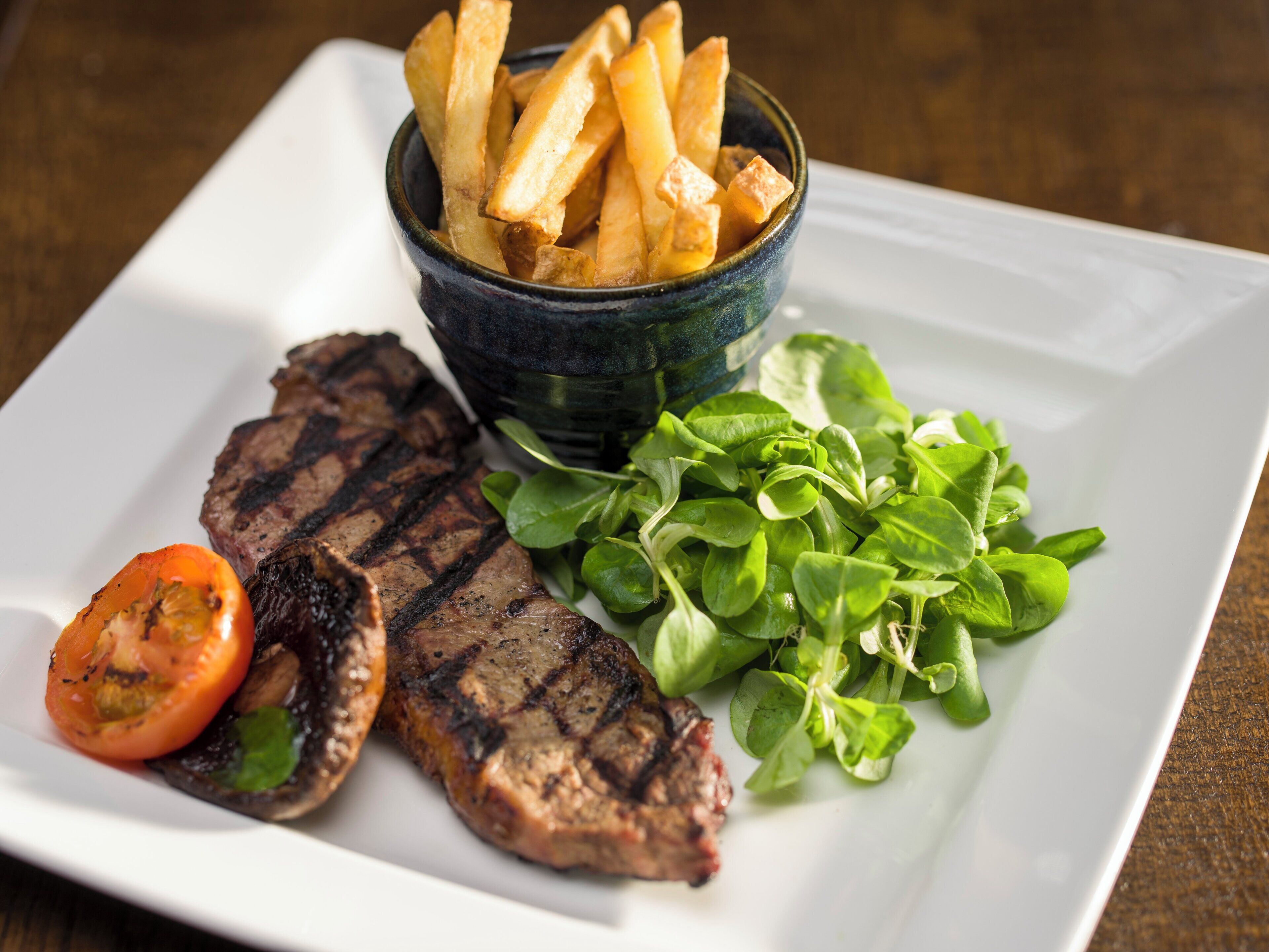 A plate with grilled steak, a grilled tomato, a side of fries, and a small bowl of greens.