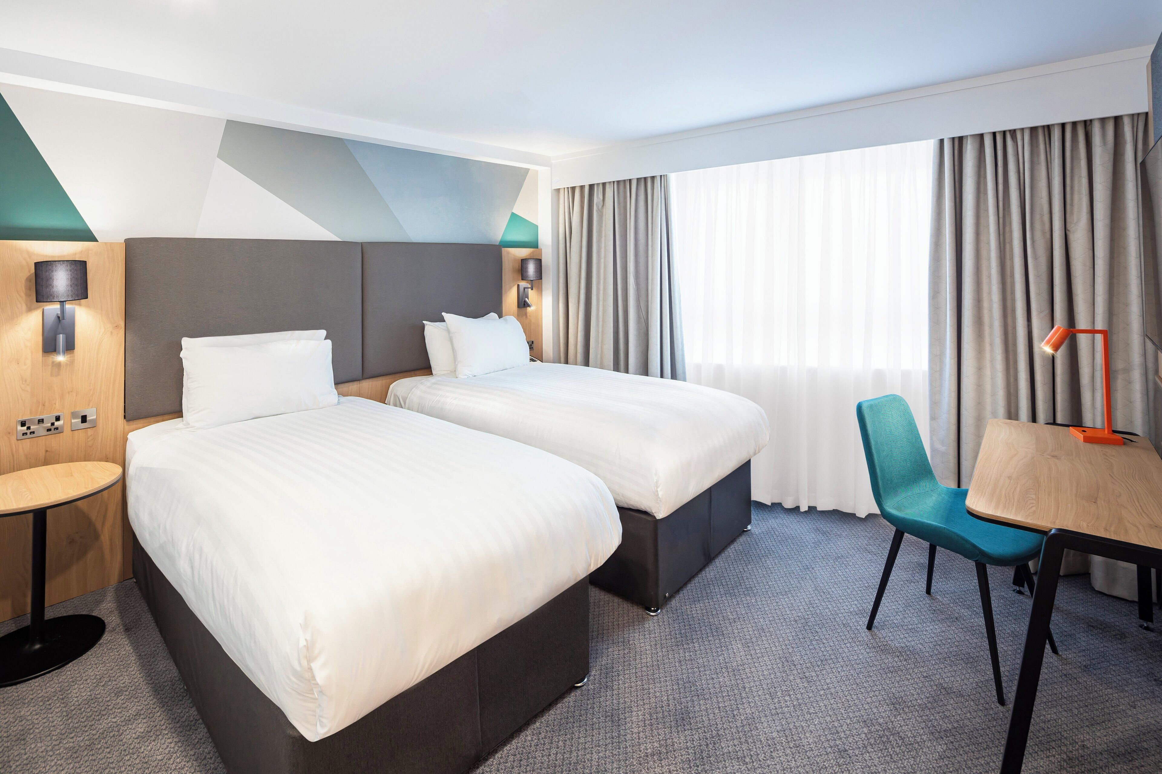 A hotel room with two beds, a desk, a chair, and a window with curtains.