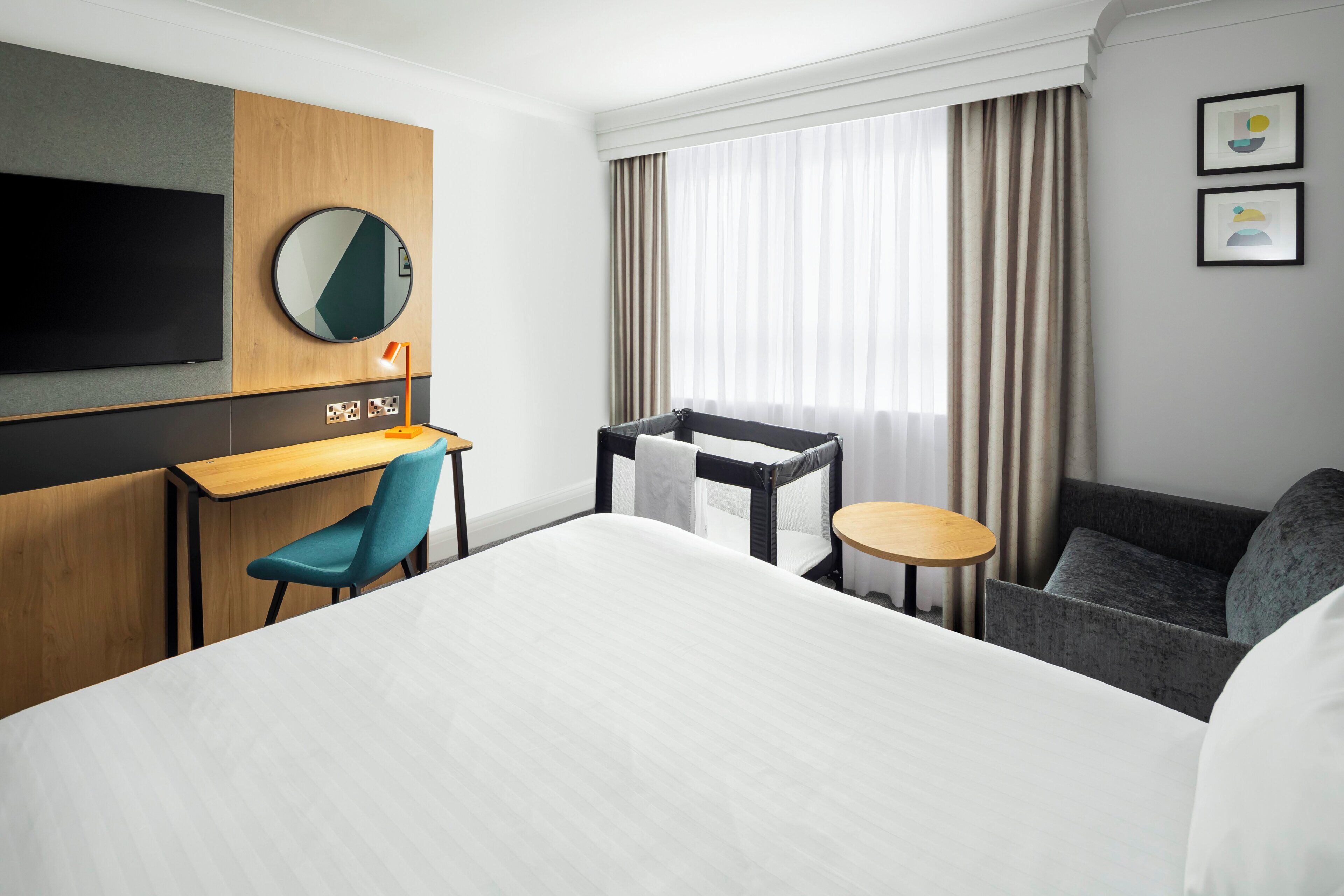 A hotel room with a bed, a desk, a chair, a small table, a TV, and a window with curtains.