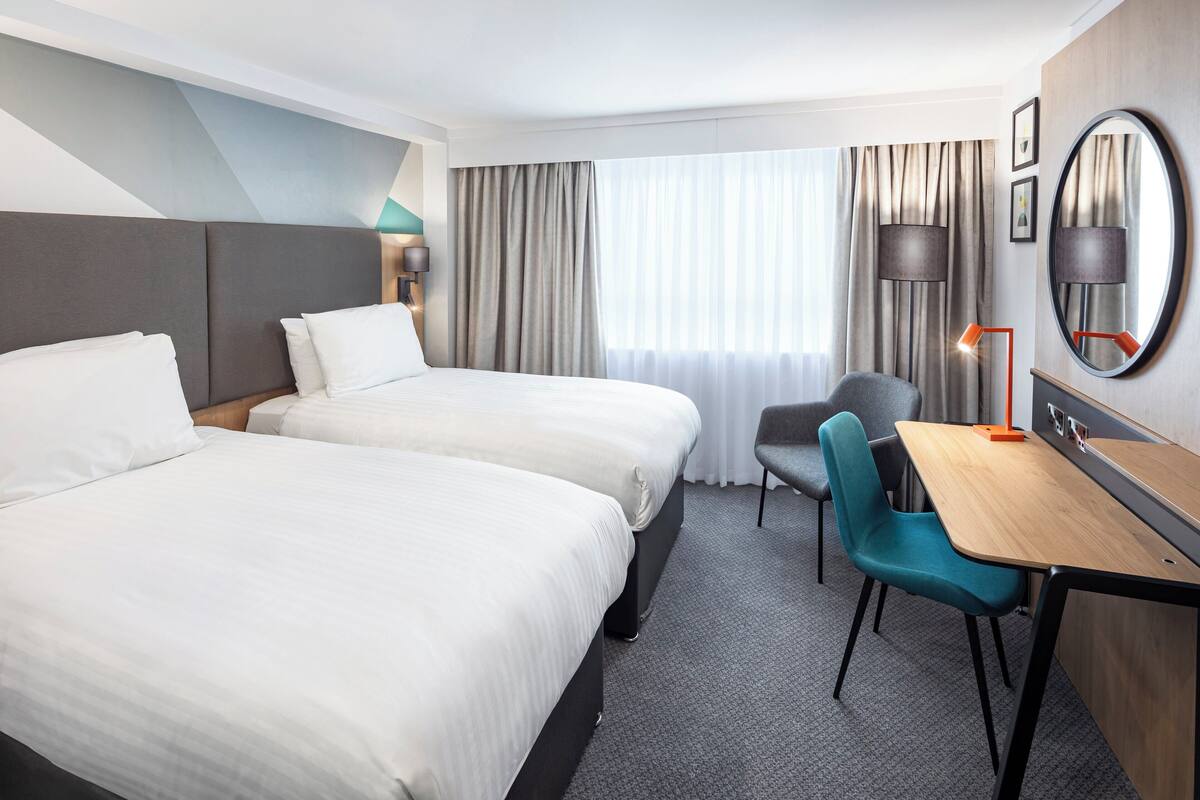 Une chambre d’hôtel avec deux lits, un bureau, une chaise, une lampe et un grand miroir.