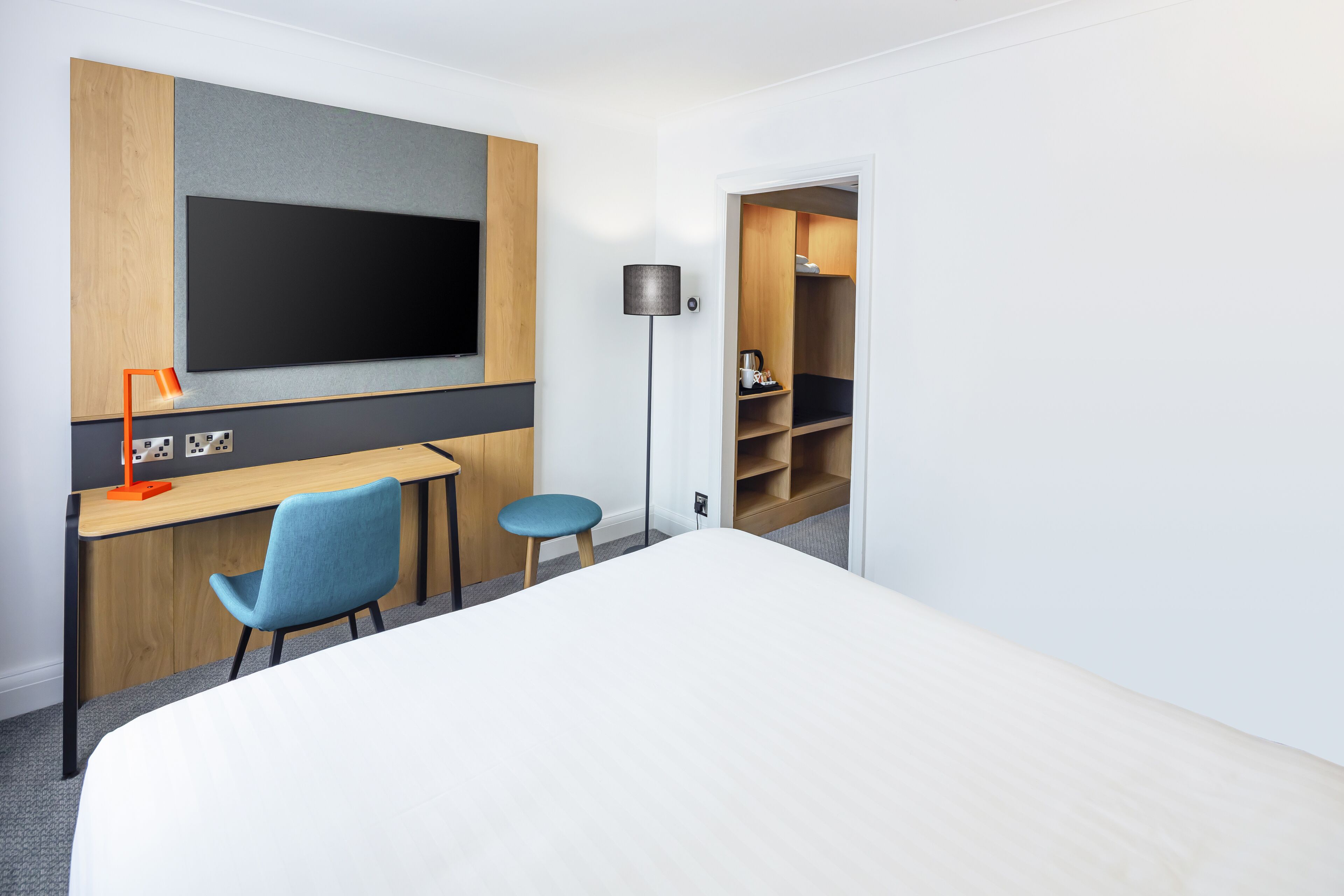 Una habitación de hotel con cama, escritorio, silla, televisor, lámpara y armario.