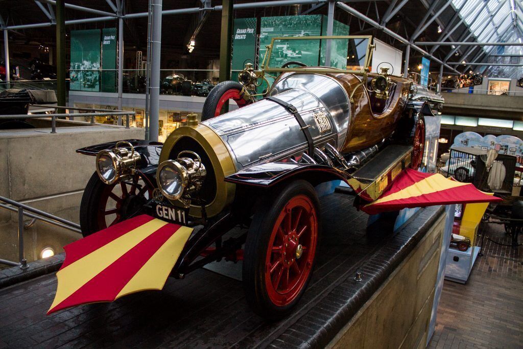 A vintage car with red wheels and a distinctive front design, displayed in an indoor exhibit with other vehicles.