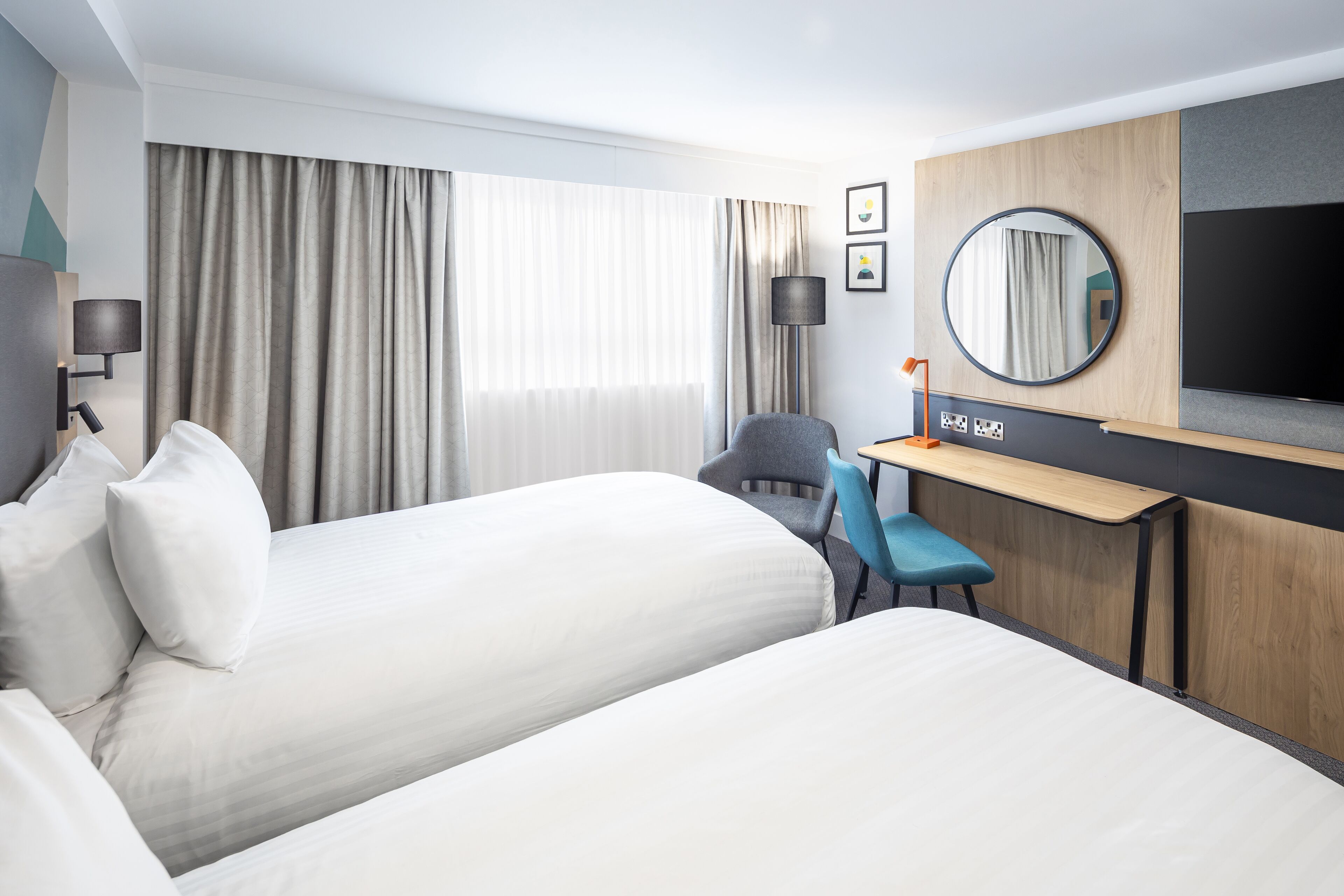 Habitación de hotel con dos camas, un escritorio, una silla, un televisor y un amplio ventanal con cortinas.