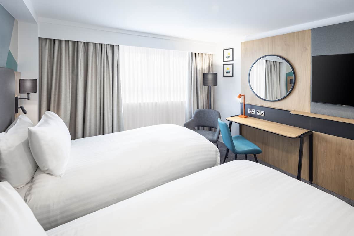 Habitación de hotel con dos camas, un escritorio, una silla, un televisor y un amplio ventanal con cortinas.