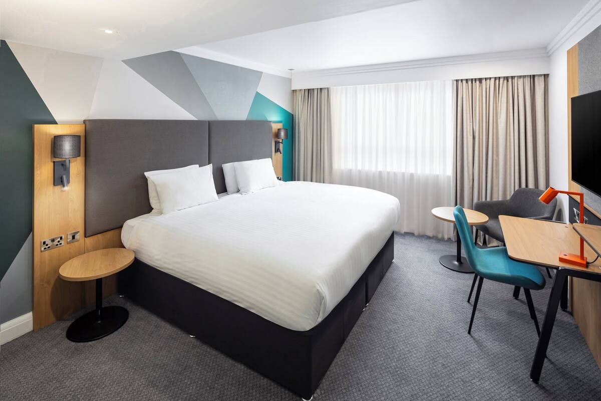 Une chambre d’hôtel moderne, dotée d’un grand lit, d’un bureau avec une chaise, d’une télévision et d’une fenêtre avec des rideaux.