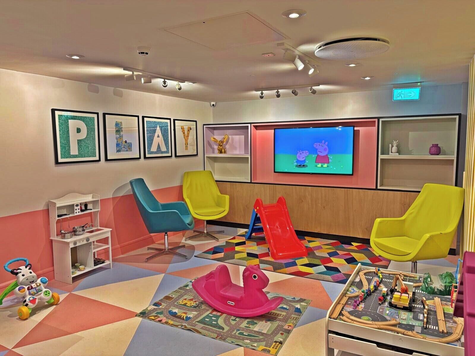 A children's play area with colorful chairs, a slide, a play mat, and a television screen displaying animated characters.