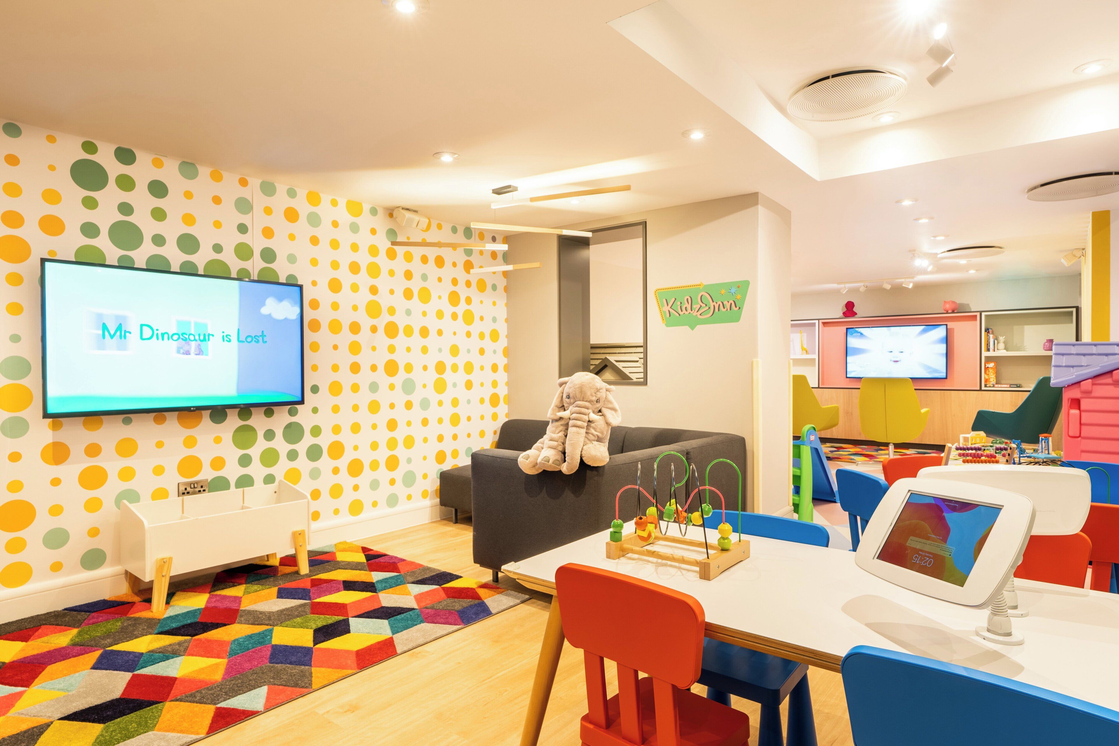 Ein Kinderspielzimmer mit einer farbenfrohen Wand mit Punkten, einem Fernseher, der eine Kindersendung zeigt, und verschiedenen Spielgeräten.