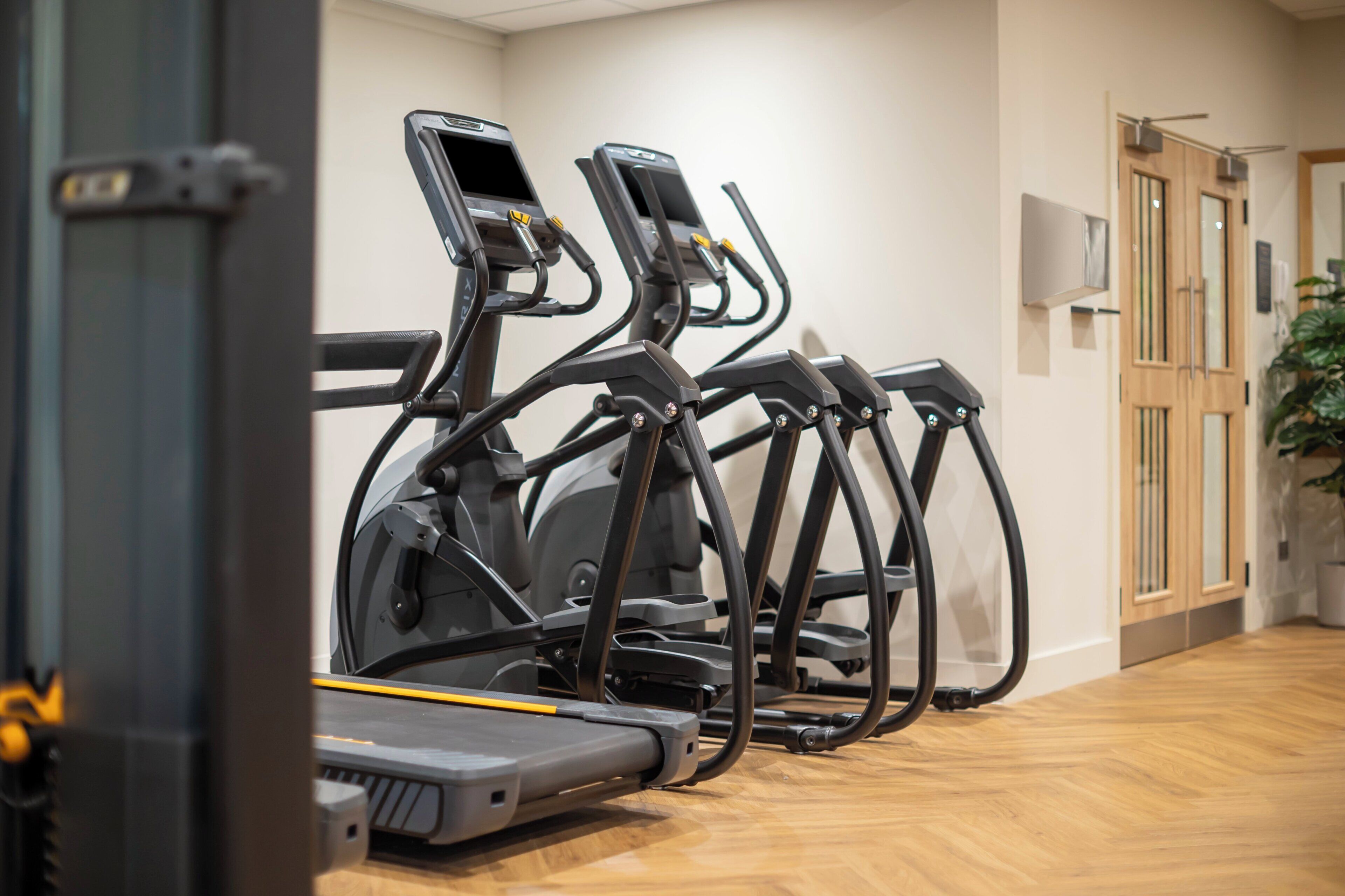 A row of stationary exercise bikes in a room with wooden flooring and a door in the background.