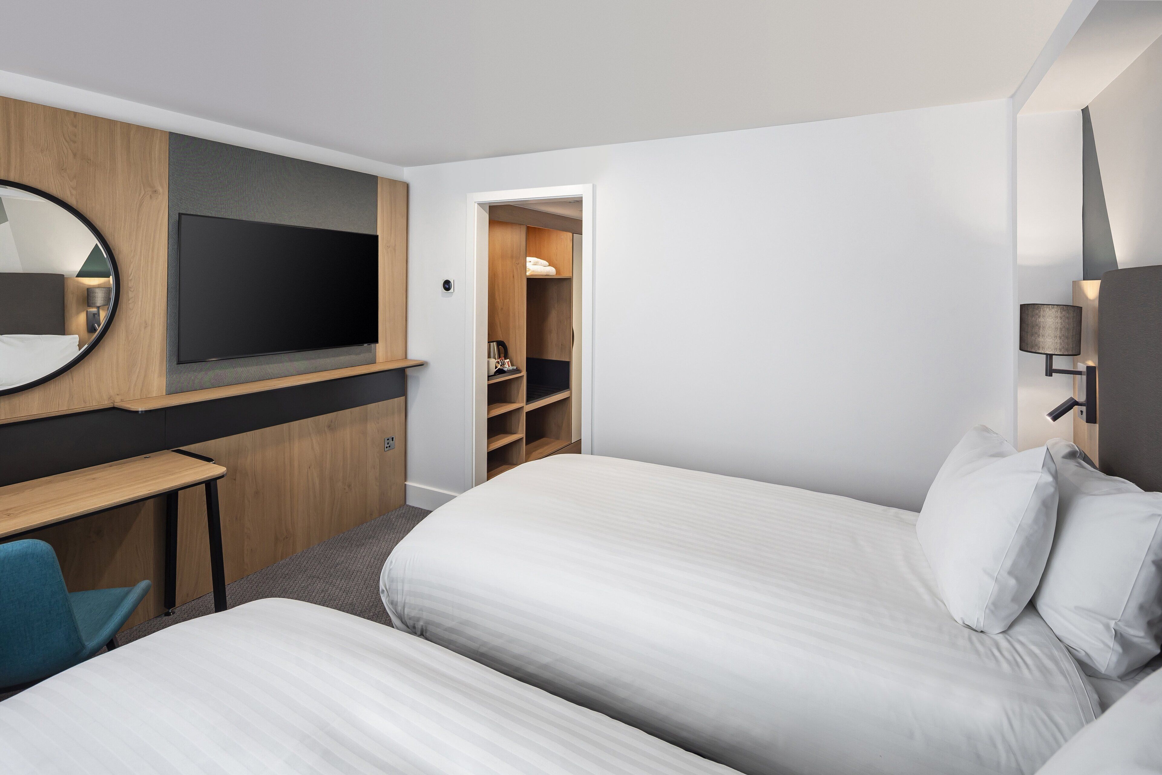 A modern hotel room with two beds, a desk, a TV, and a walk-in closet.