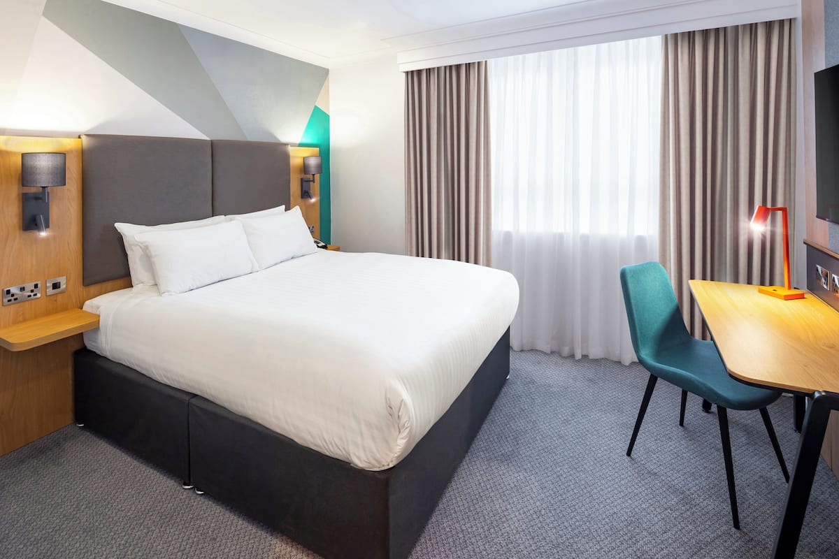 Une chambre d’hôtel avec un grand lit, un bureau avec une chaise, une télévision et une fenêtre avec des rideaux.