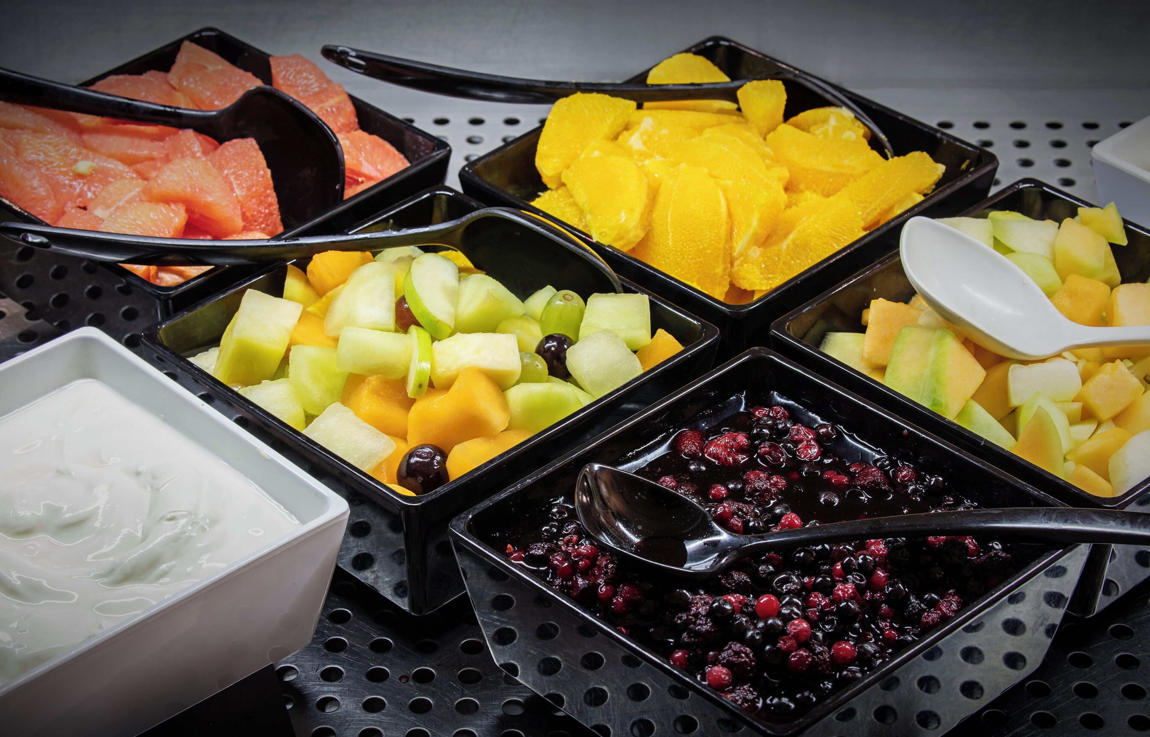 Diversos cuencos de fruta con cucharas, dispuestos sobre una bandeja perforada.