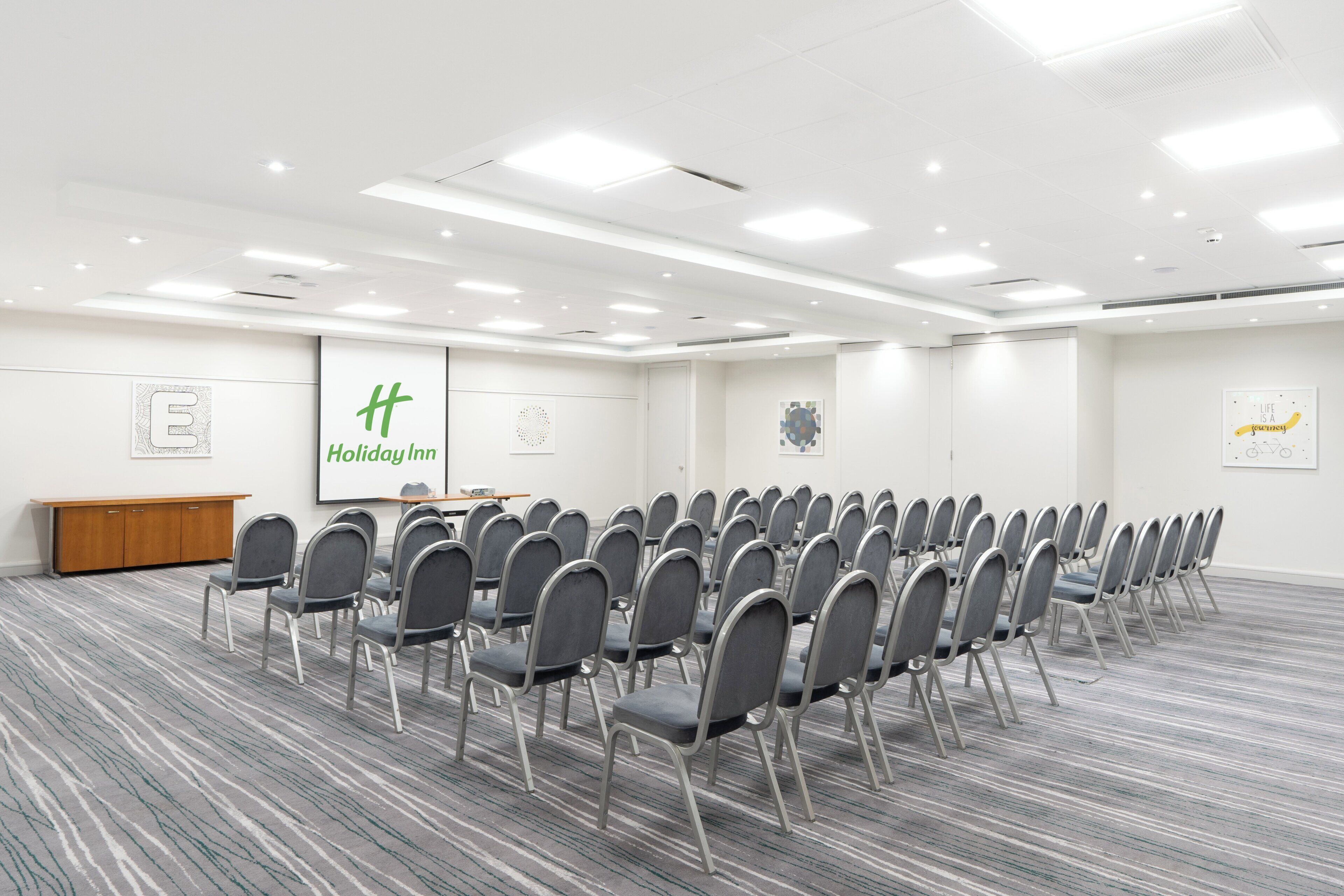Un salón de conferencias con filas de sillas, un atril y un gran letrero de 'Holiday Inn' en la pared.