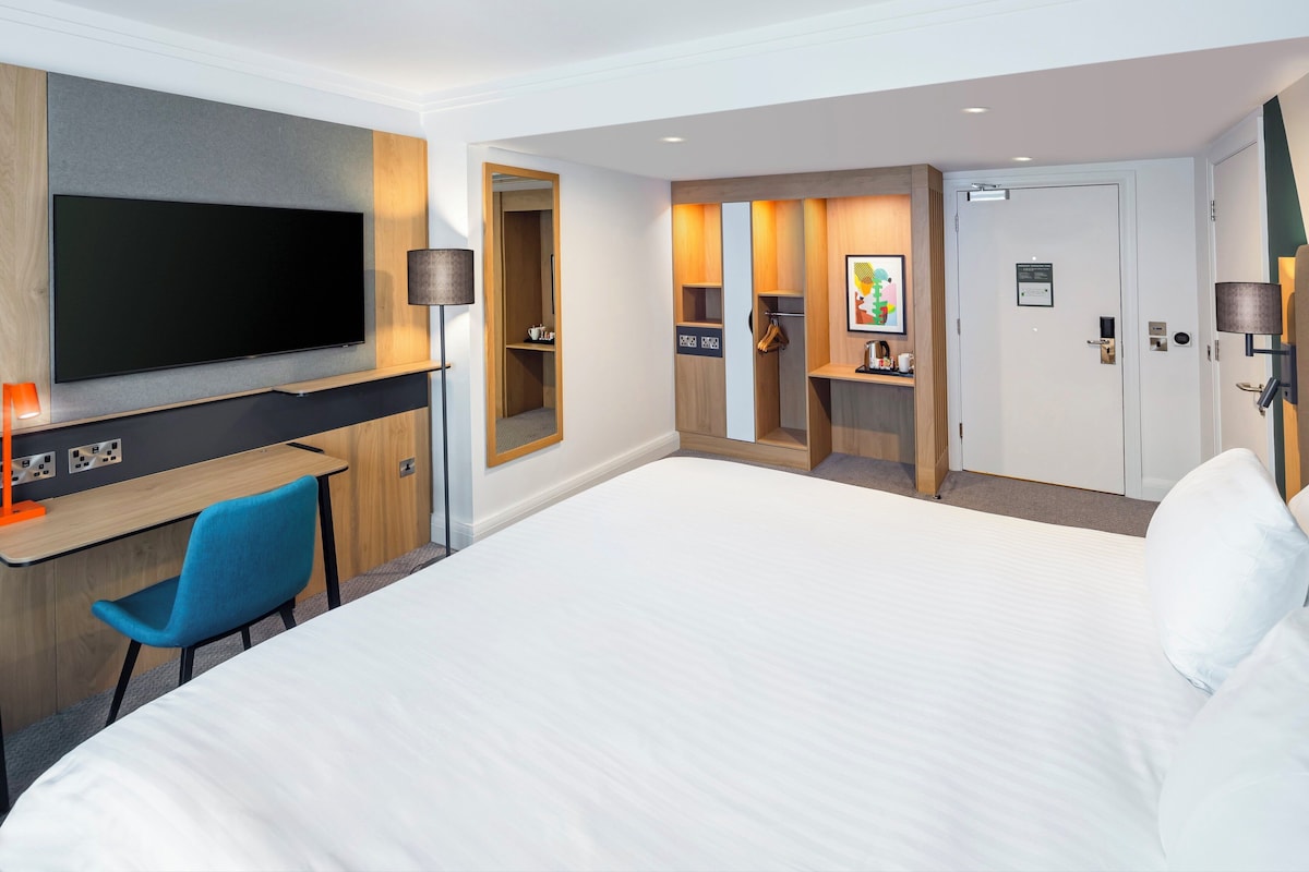 A hotel room with a large bed, a desk with a flat-screen TV, a blue chair, a wardrobe, and a door with a sign.
